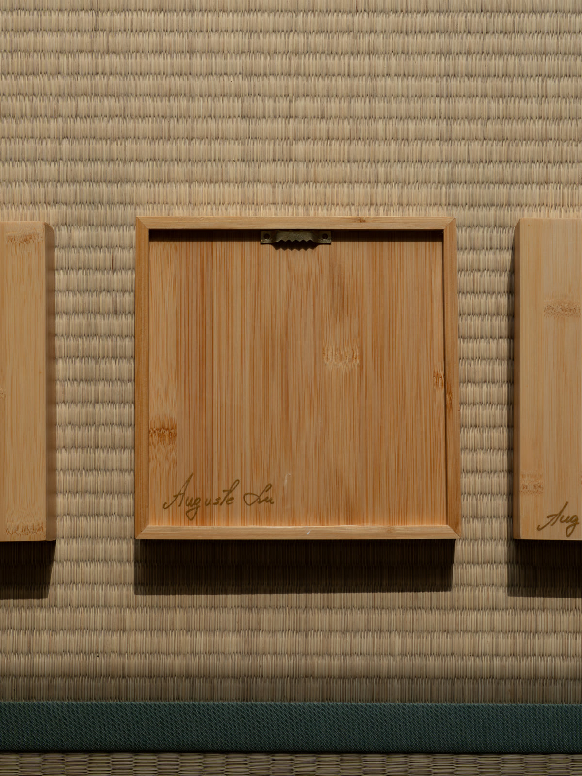 A square light wood frame with a hinged lid, plced on a tatami mat. The lid is engraved with the name 'Auguste La' in cursive script.